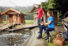 A man in a red shirt and a woman in a blue shirt fish by a lake near wooden cabins. The scene features camping gear.
