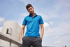 A man wearing a blue mercerised polo shirt stands outdoors, hands in pockets, against a cloudy sky backdrop.
