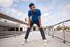 A man in a blue polo shirt and grey trousers stands outdoors, leaning forward with hands on knees against a modern backdrop.