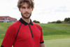 A man in a red zip polo shirt with black accents stands on a golf course background. The shirt features a collar and a logo.