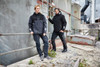 A padded hardshell workwear jacket in black and grey worn by two individuals posing against an industrial backdrop.