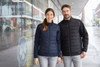 A navy and black soft shell jacket is worn by a woman and a man in an urban setting. Both jackets have a logo.