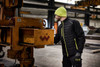 A men's hybrid jacket in black with bright yellow accents, worn by a man in a beanie and gloves near industrial equipment.