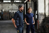 A man and woman stand in an industrial setting, wearing dark blue and navy work polo shirts and matching work pants.
