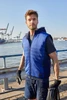 A blue padded hybrid vest with a hood, worn by a man by the waterfront, featuring a logo on the front.