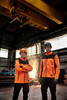 Two individuals wearing bright orange softshell jackets in a workshop setting, with industrial equipment and sparks in the background.