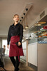 A woman wears a burgundy apron with a black shirt, standing in a stylish indoor setting with modern lighting.