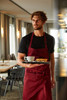 A maroon apron with bib displayed on a model holding a tray with a coffee and food in a restaurant setting.