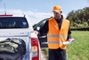 An orange safety vest with reflective stripes worn by a man near a vehicle, surrounded by greenery.