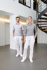 A man and woman stand in a modern indoor space wearing grey tops and white comfort pants. The setting features a spiral staircase.