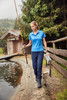 A woman in a blue polo shirt walks by a pond, holding a fishing rod and carrying a fishing tackle box, surrounded by nature.