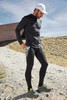 A man in a black zip hoodie and black leggings stands outdoors, wearing a white cap and sporty shoes. The background features rocky terrain.