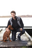 A man in a black fleece jacket kneels beside a brown dog on a wooden dock by a lake. The scene is outdoor and sunny.