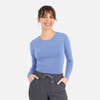 A woman is wearing a long-sleeve, light blue underscrub tee paired with grey pants. The shirt has a logo.