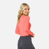 A woman models a long-sleeve, coral underscrub tee, paired with dark grey pants. The shirt features a branding logo.