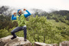 A man wearing a green and blue knitted fleece vest poses on a rock outdoors, surrounded by greenery and mountains.