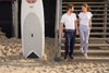 A pair of ladies jog-pants in grey, featured alongside a man and woman on a sandy beach near steps and a surfboard.