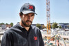 A man wears a black polo shirt and matching cap, featuring a logo, against a construction site backdrop.