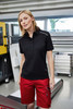 A woman wearing a black polo shirt and red bermuda shorts, standing in an industrial workspace.