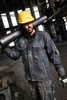 A man in a black softshell jacket carries a pipe over his shoulder, wearing a yellow hard hat and gloves.