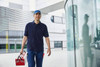 A man wearing a navy polo shirt and cap carries a toolbox while walking past a glass building.