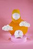 A white kids' trucker cap with a logo, displayed with yellow and pink geometric shapes and fluffy clouds.