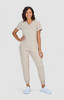 A woman stands smiling in beige scrubs and white sneakers. The outfit features a top with a v-neck and pants with pockets.