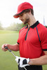 A red 5 panel sports cap worn by a man in a red polo shirt, holding a golf club on a green. The cap features a logo.