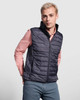 A man wearing a navy padded vest over a light pink shirt, standing against a grey background. The vest has a logo.