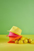 A yellow, pink, and orange hat stacked on a yellow surface, with lemons beside them against a green background.