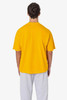 A man stands with his back to the camera, wearing a bright yellow short sleeve pocket tee and grey sweatpants.
