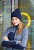 A woman wearing a navy high brim beanie and a grey zip-up jacket stands with her arms crossed. The background features a blurred circle of light.
