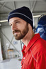 A reflective beanie in navy blue, worn by a man with a beard, inside a hangar or workshop setting.