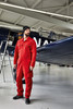 A man in a red jumpsuit and black beanie stands next to an airplane in a hangar. The image features a logo on the beanie.