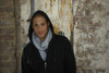 A person wearing a grey fleece scarf and a black hoodie stands against a weathered brick wall.