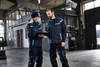 A padded work vest in navy blue with teal accents, worn by two people in a workshop setting, one with a clipboard.