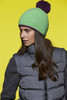 A green knitted hat with a purple pompom, worn by a woman with long hair, against a bright yellow background.
