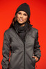 A woman wears a ribbed black scarf and matching beanie, paired with a grey jacket against a vibrant orange background.
