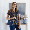 A smiling woman wearing a floral navy top holds a small navy bag, standing beside a decorative vase and artwork.