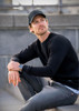 A black sweatshirt with long sleeves worn by a man sitting on stairs, paired with grey jeans and a cap.