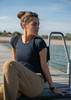 A woman sits on a dock wearing a navy blue T-shirt and beige trousers, with a serene water background.