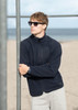 A man wearing a navy polar fleece jacket and sunglasses stands by a beach, leaning against a metal pole.