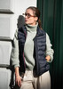 A navy vest is worn over a light green sweater, paired with cream trousers. The model has glasses and is leaning against a wall.