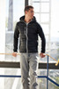A man is standing inside near a window wearing a black hybrid jacket with a hood, featuring a zip front and a logo.