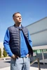 A padded navy vest worn over a blue jumper, modelled by a man standing outdoors under a clear sky.
