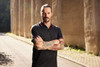 A man with tattoos is wearing a black polo shirt with a contrasting collar, standing against a textured background.