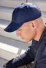 A navy blue 6 panel cap is worn by a man with a shaved head, sitting on stairs. The cap features a logo.