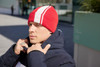 A red winter beanie with white stripes, worn by a person in a dark jacket, set against an outdoor background.