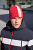 A red winter beanie with white stripes, worn by a person in a navy jacket. The background shows an outdoor setting.