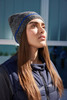 A coarse knitted beanie featuring grey and blue shades, worn by a model with long hair, against a modern backdrop.